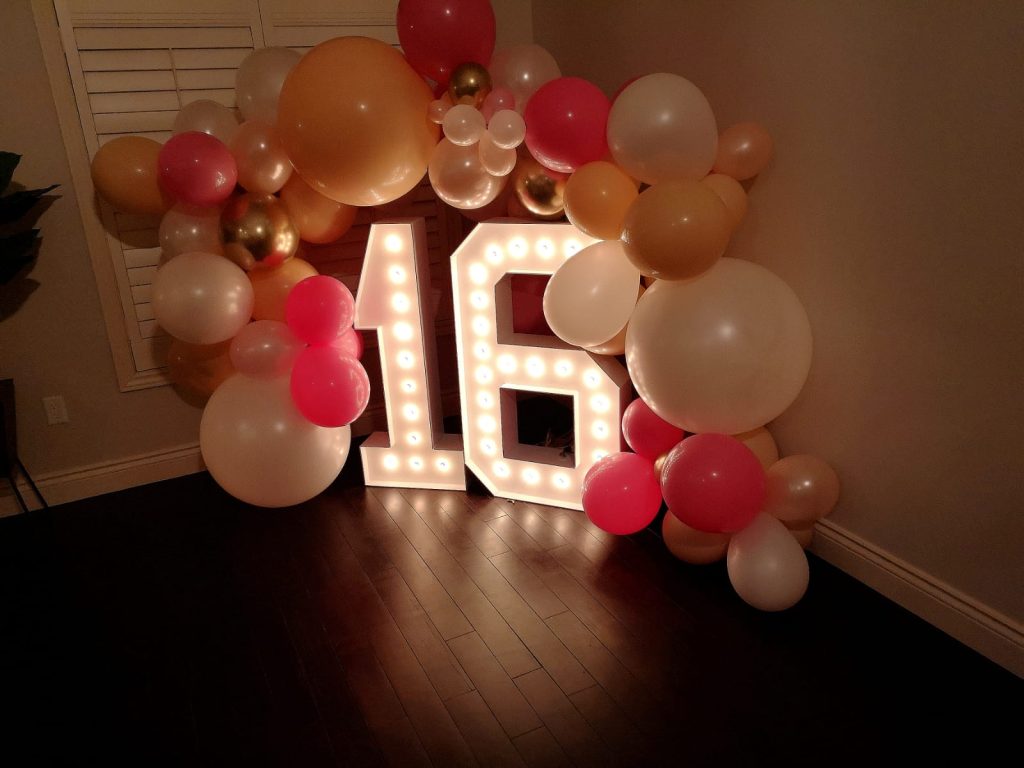 Gravenhurst balloon garland in soft party colors frames a birthday photo area with glowing marquee numbers spelling the child’s age, set up for guest photos.